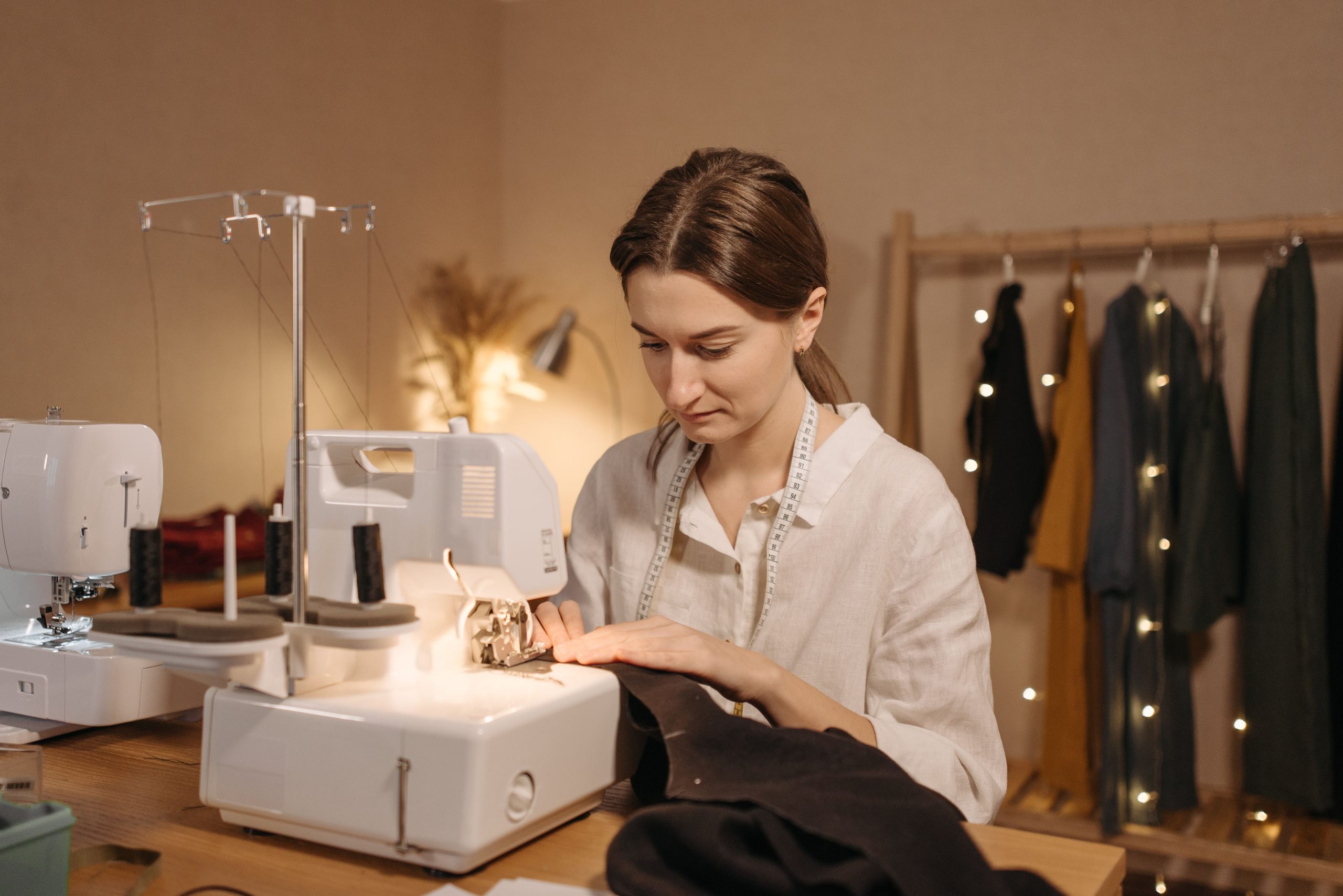 A woman using a sewing machine in a cozy workshop with clothes hanging and string lights.
